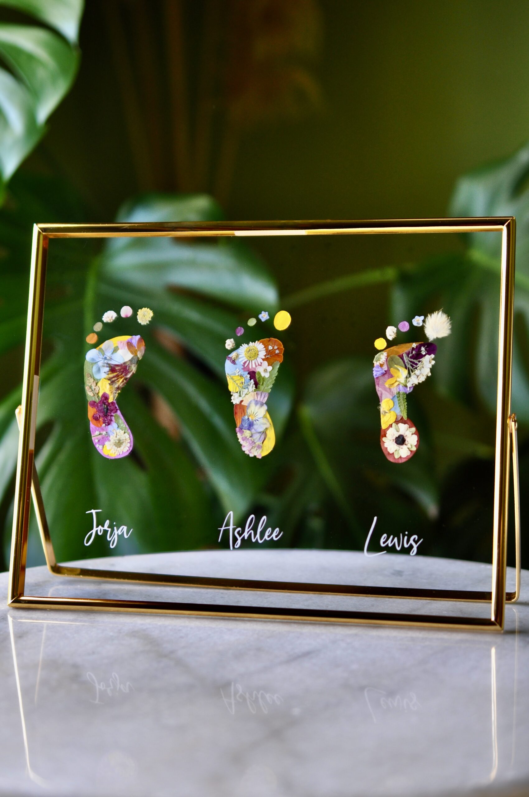 Three colourful baby footprints made from pressed flowers with names Jorja, Ashlee, and Lewis in a gold frame.