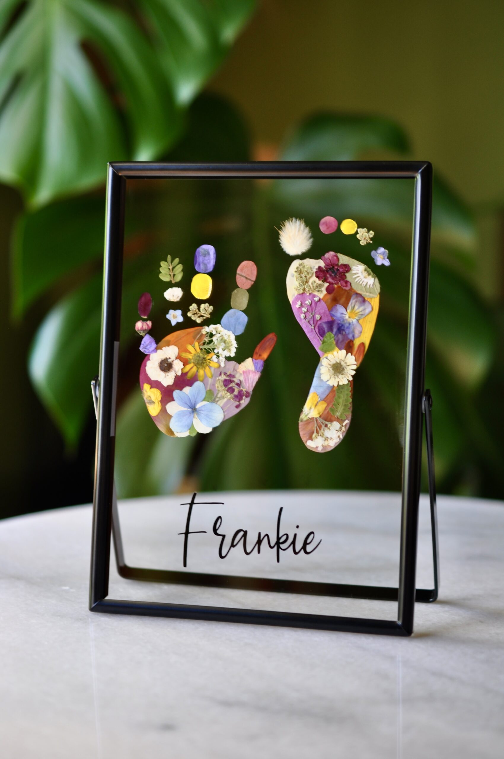Custom baby hand and footprint keepsake made from real pressed flowers, labelled with baby's name and birth date in a black double-glass frame