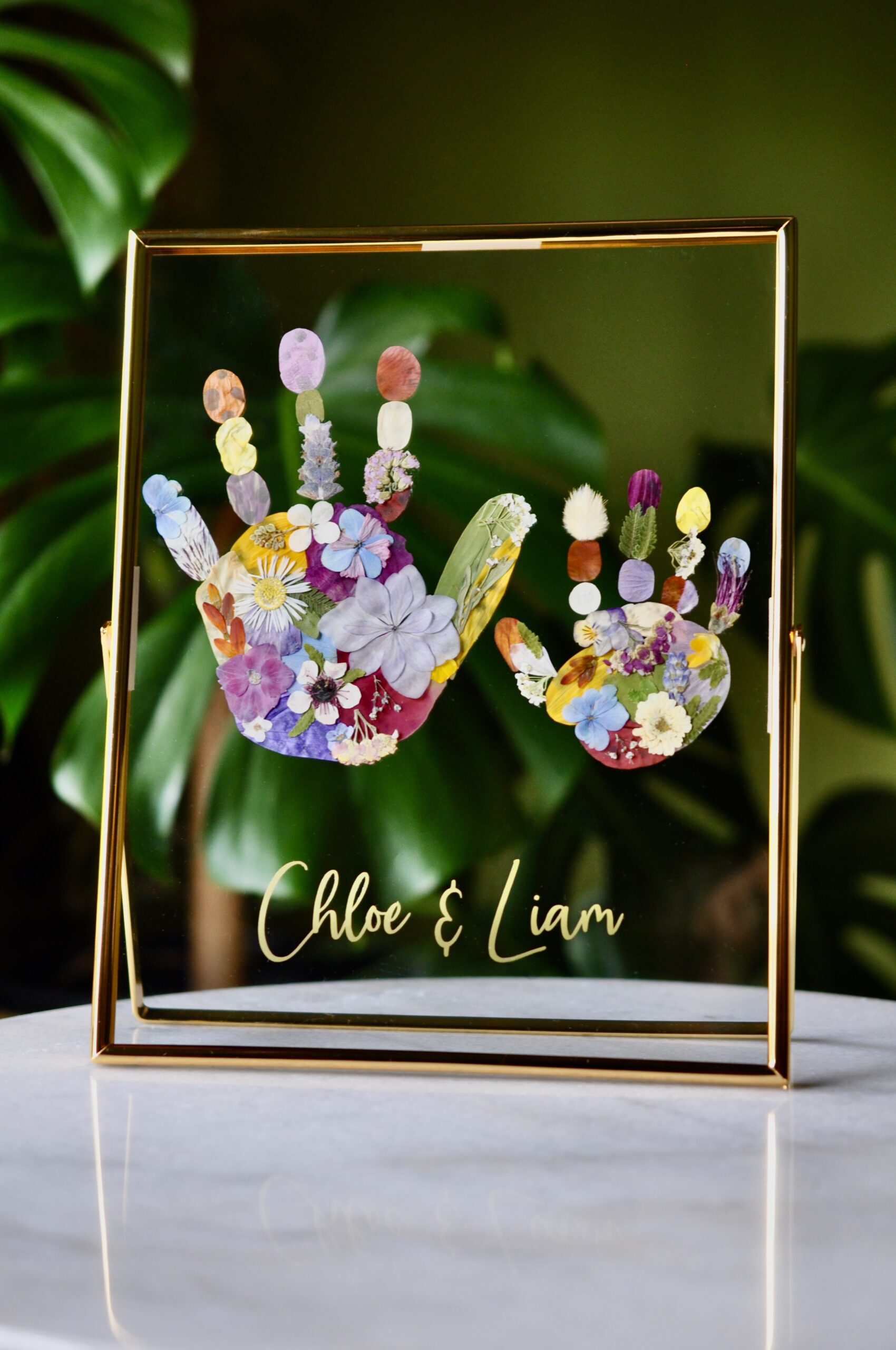 Framed sibling handprints made from real pressed flowers with the names Chloe and Liam, set in gold glass frame.