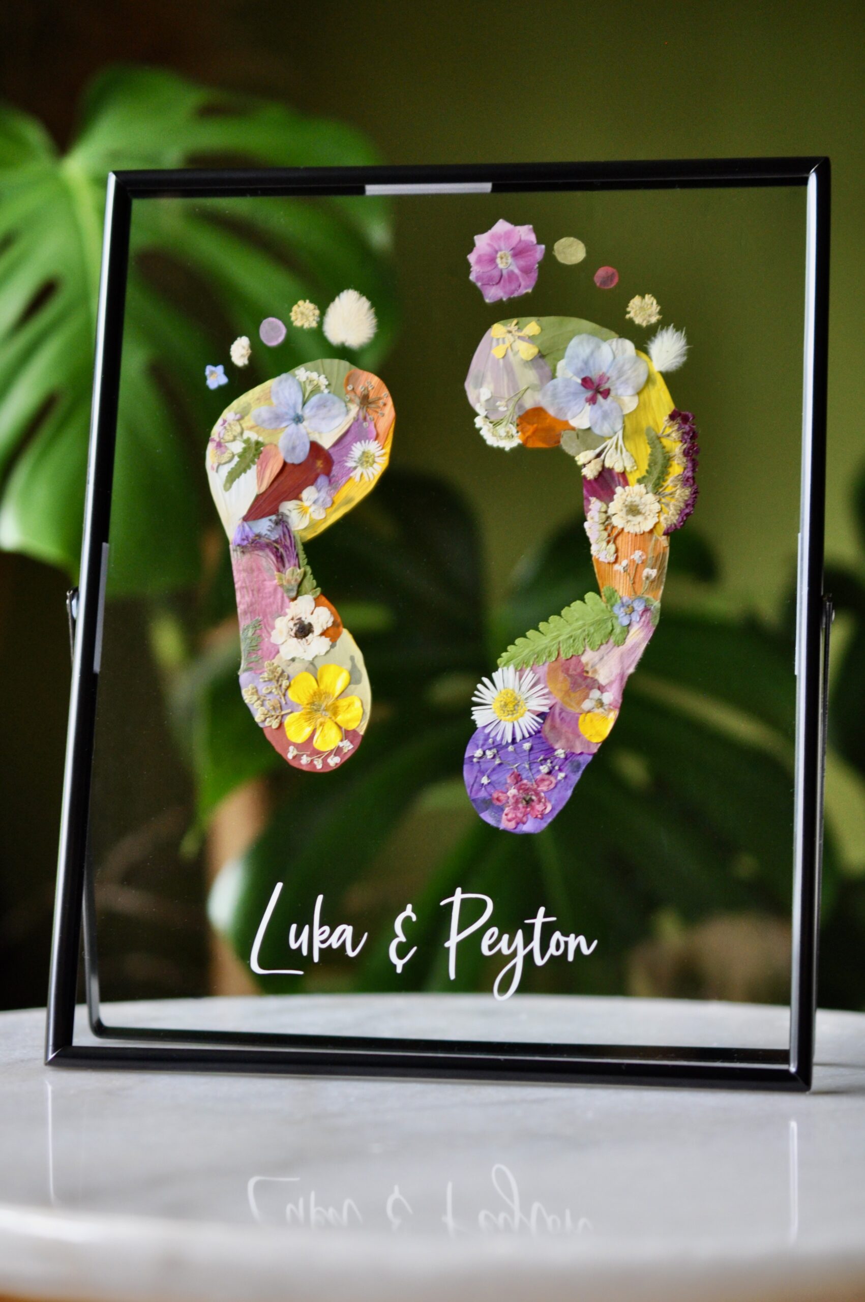Sibling baby footprints made from real pressed flowers in a double glass frame with names "Luka & Peyton" beneath.