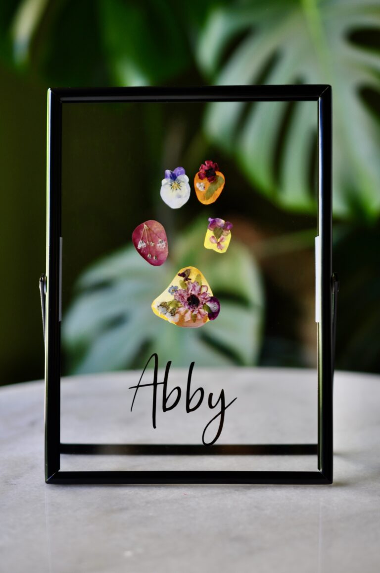 Dog paw print made from real pressed flowers in a black glass frame with the name “Abby.”