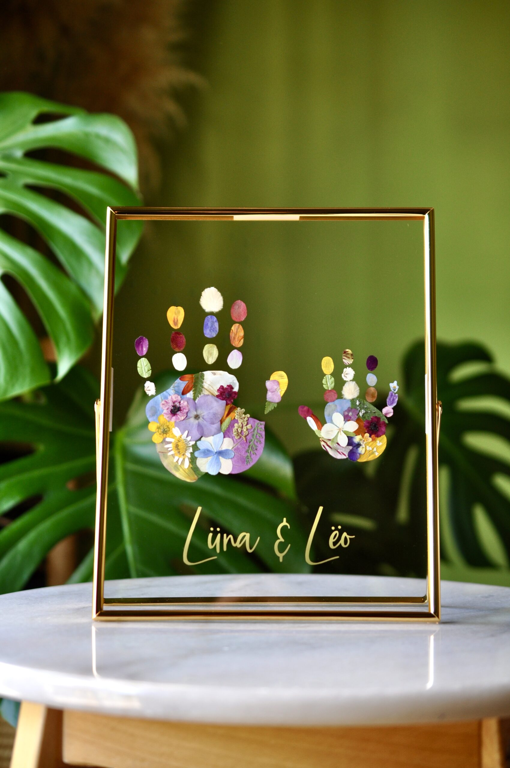 Framed sibling handprints made from real pressed flowers with the names Lüna and Leo, set in gold glass frame.