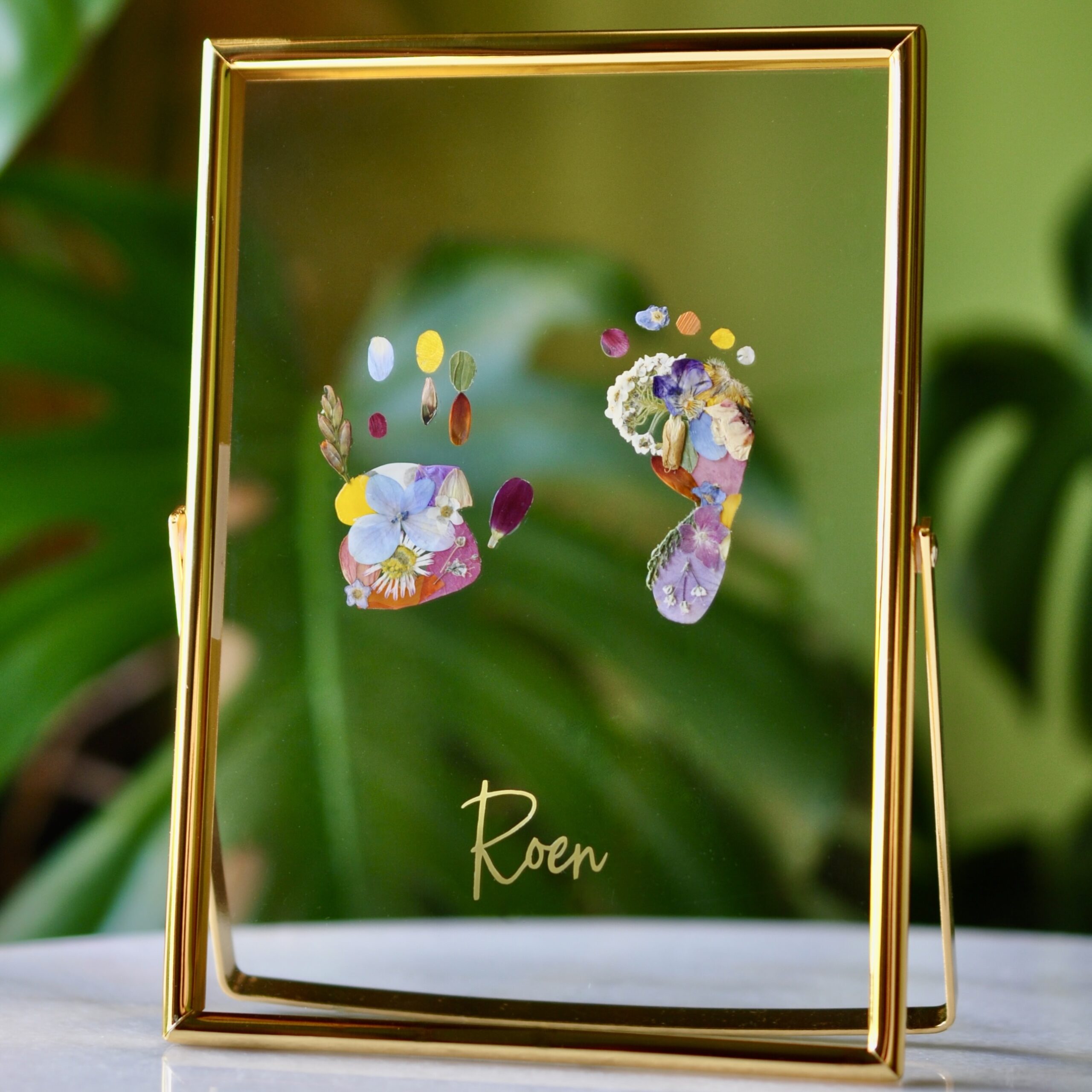 Framed baby footprints made from real pressed flowers, displayed in a gold double glass frame with the name “Roen” in gold script