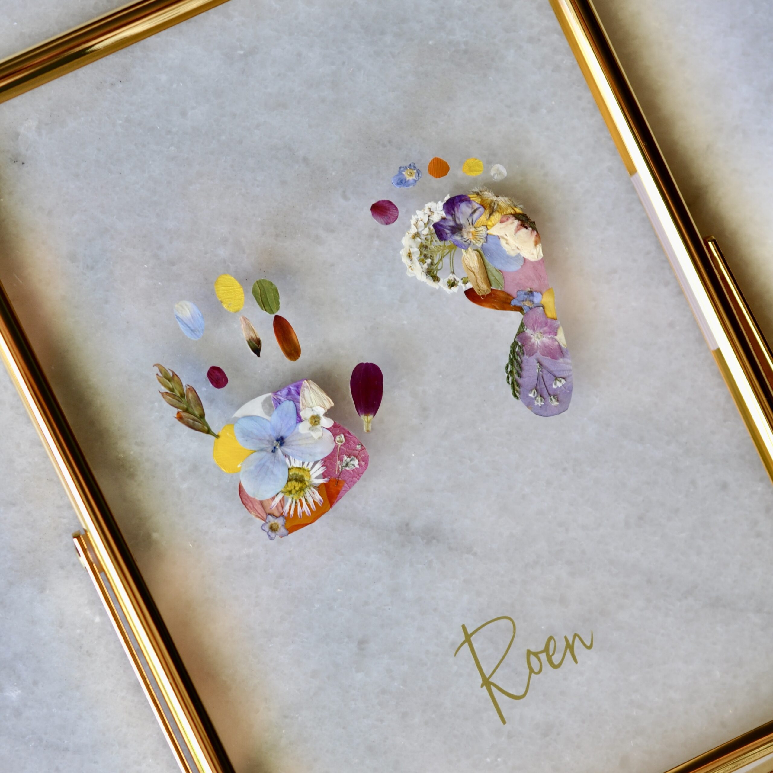 Framed baby footprints made from real pressed flowers, displayed in a gold double glass frame with the name “Roen” in gold script