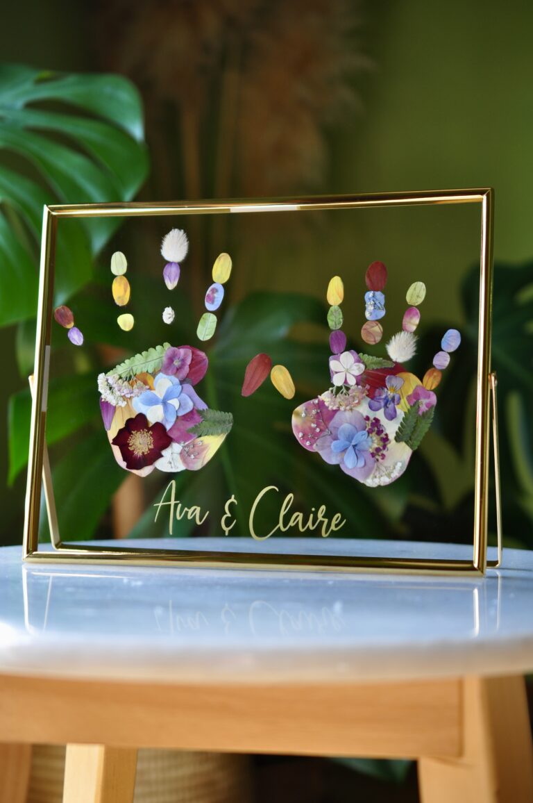 Framed sibling handprints made from real pressed flowers with the names Ava and Chloe, set in gold glass frame.